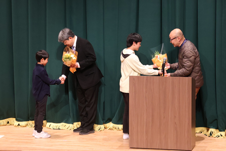 児童から花束贈呈。 ※人事異動で初等学校を離れる事務職員の鈴木さんが欠席のため、代わりに加藤事務長が受け取りました
